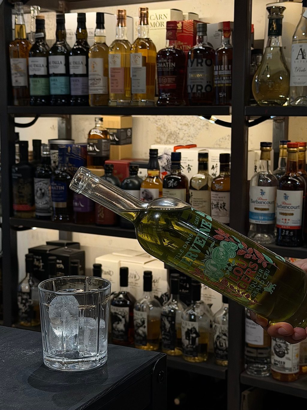 Service de Lou Gers Verveine, bouteille en main devant des étagères de spiritueux
